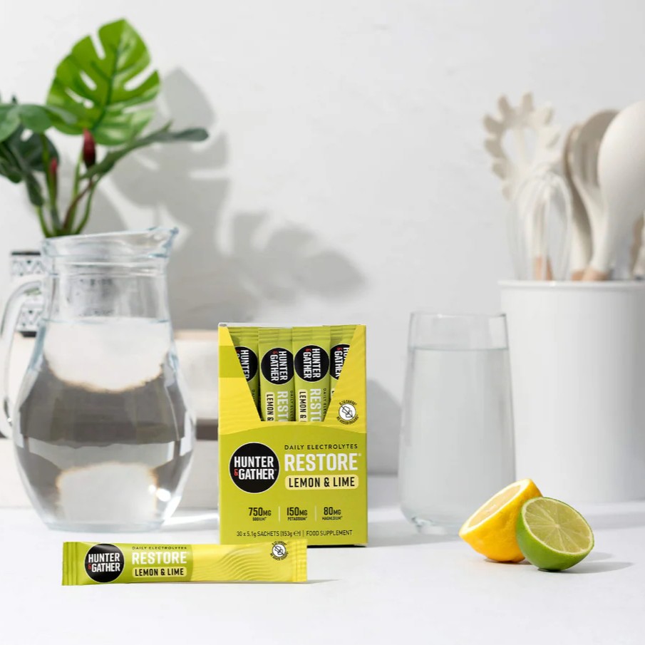 RESTORE Lemon & Lime box and sachet beside a glass and jug of water with fresh lemon and lime on a kitchen counter.